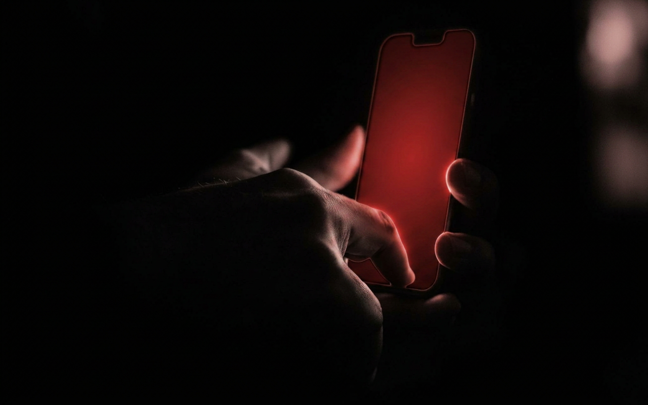 Editorial-Schwarz-Weiß-Foto einer erwachsenen Hand, die im Dunkeln nach einem leuchtenden Smartphone greift, das Bildschirmlicht fängt sich in den Fingerspitzen